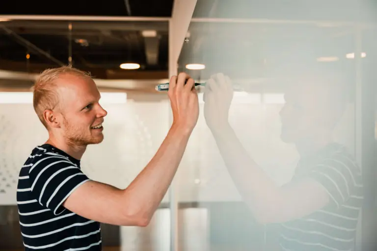 Een glimlachende man met kort blond haar, gekleed in een gestreept blauw-wit T-shirt, schrijft geconcentreerd met een whiteboard-marker op een glazen whiteboard. Zijn reflectie is zichtbaar op het glanzende oppervlak. De omgeving is licht en modern, met plafondverlichting die de kamer een warme gloed geeft.