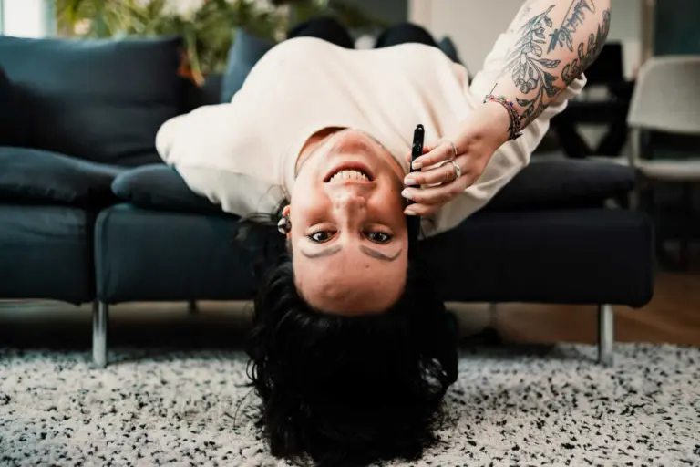 An upside-down view of a young woman lying on her back on a speckled gray and white carpet. She is holding a smartphone in her hand, her dark curly hair sprawled on the floor, and sporting a tattooed arm. She is smiling broadly, wearing a cream-colored sweater and has a stud earring in her ear. In the background, there is a dark blue sofa with cushions and a potted plant, suggesting a cozy living room setting.